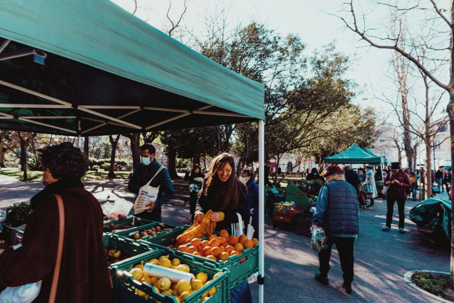People shopping for fruits and vegetables at an outdoor market under green tents on a sunny day.
