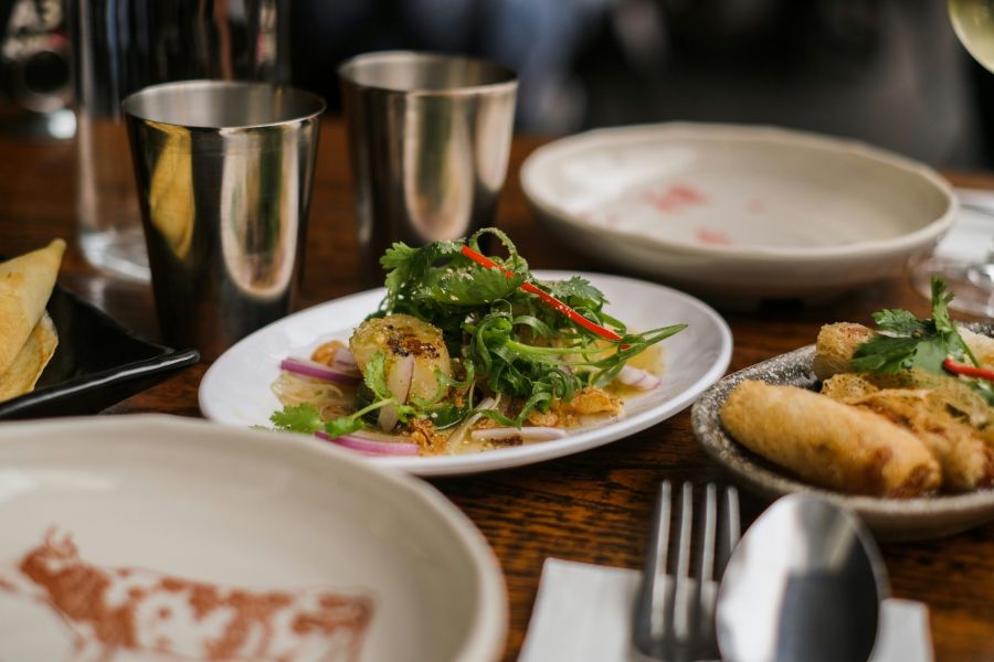 A plate of salad with greens and chili, next to metal cups and more plates of food on a wooden table.