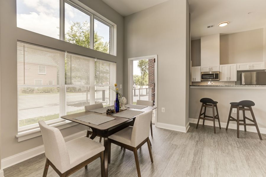 Messenger Place apartment homes with Modern dining area with four chairs, large windows, and a view of a kitchen with bar stools in the background.