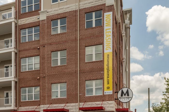 Attain on 5th apartment homes with Red brick apartment building with a large yellow "Now Leasing" banner and a Messenger Place sign.