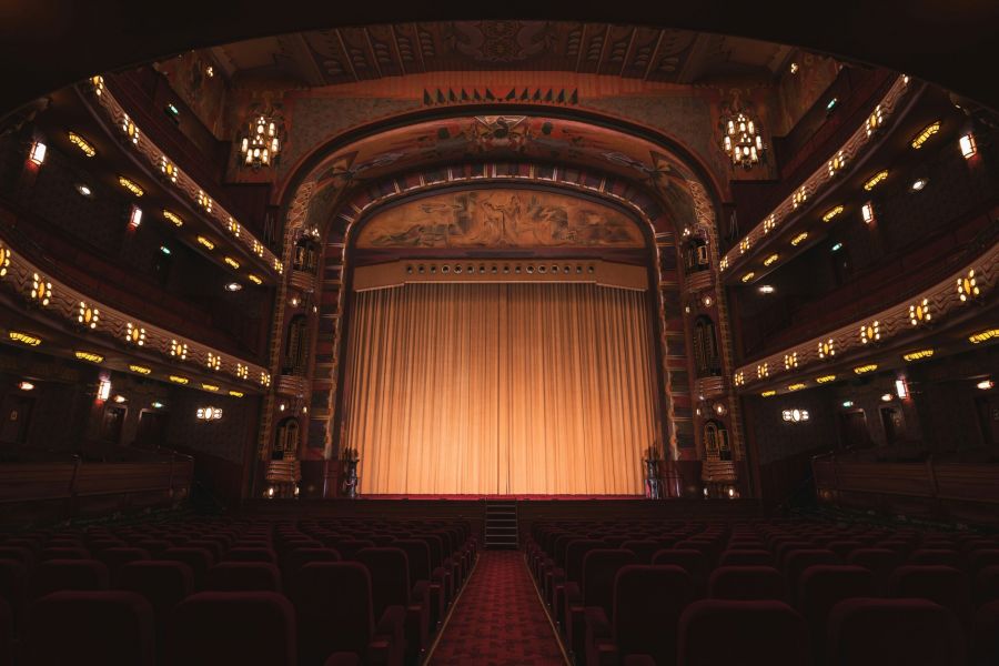 Ornate theater interior with red seats facing a large stage covered by golden curtains.